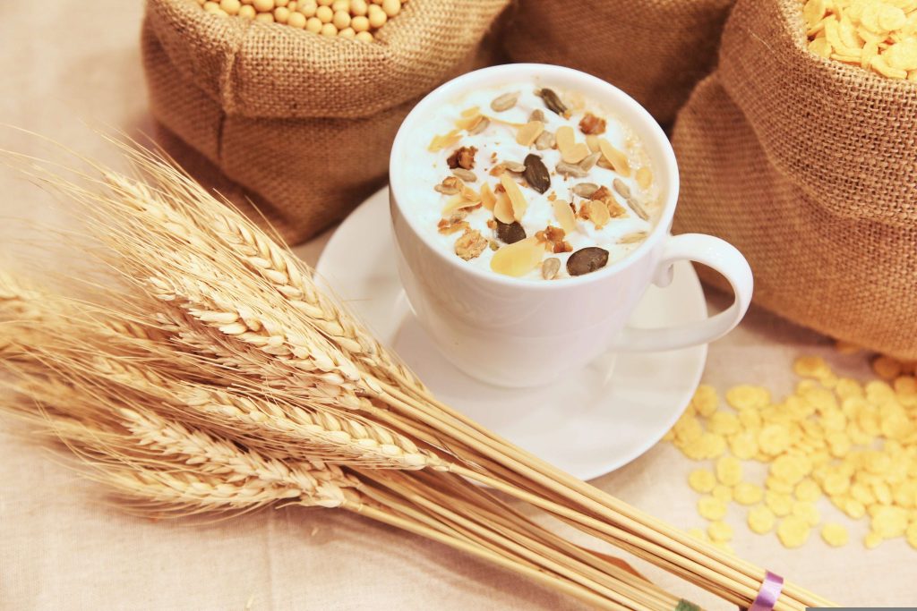 A white cup of warm cereal drink topped with nuts and seeds, surrounded by wheat stalks and grain sacks