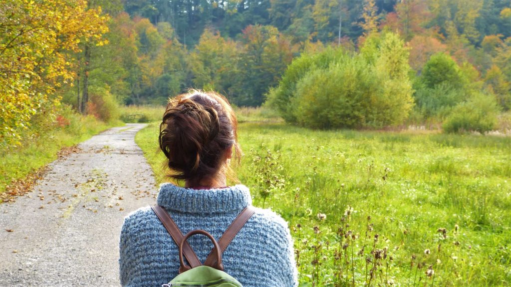 Rear view of a woman with a backpack walking along a quiet trail surrounded by green fields and trees