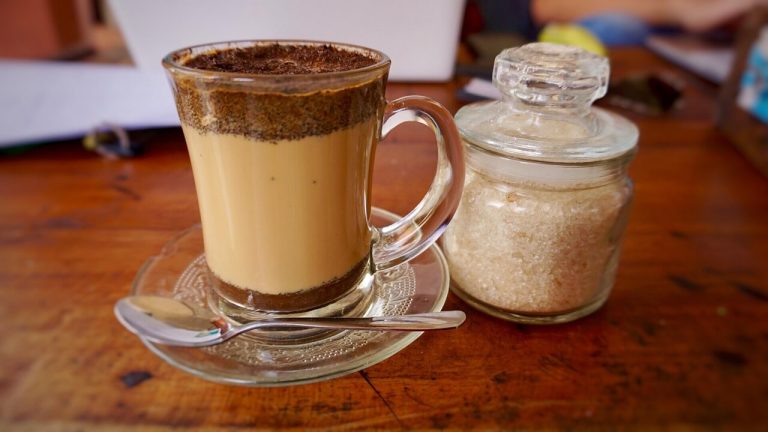 A clear glass mug filled with layered milk coffee sitting on a wooden table next to a glass jar of raw refined sugar, highlighting traditional sweetening methods.