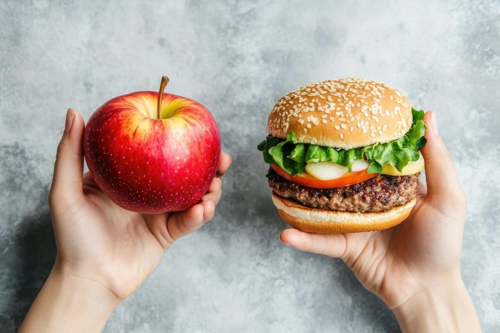 A hands hold a fresh red apple and a large cheeseburger, illustrating the healthy daily steps seniors can take to avoid ultra-processed foods.