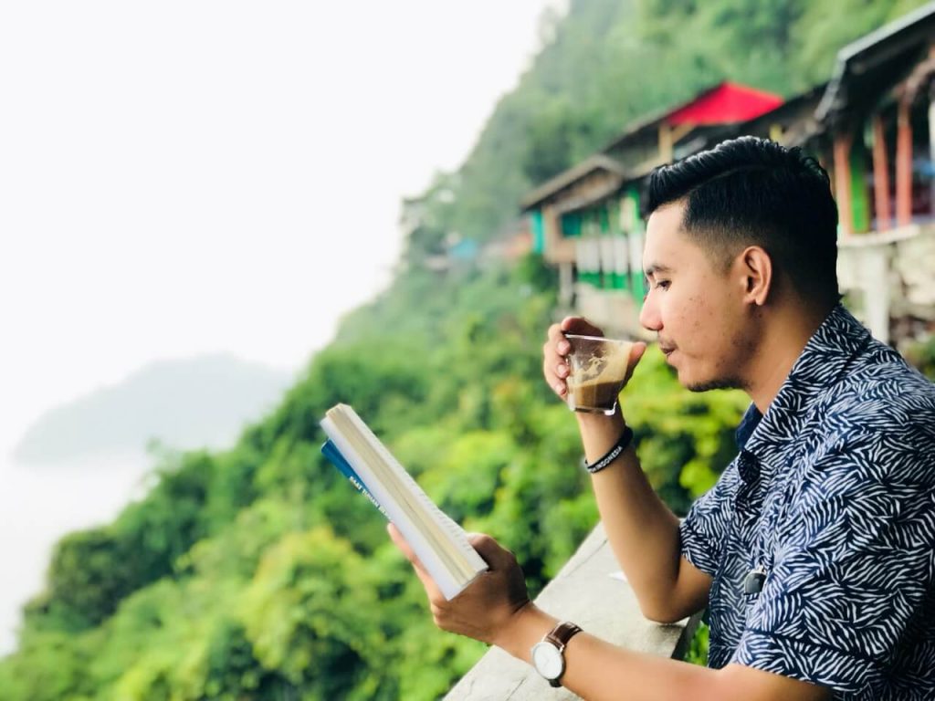 A man in a blue patterned shirt enjoying a warm cup of coffee while reading a book on a balcony overlooking lush green hills, representing a healthy morning routine.