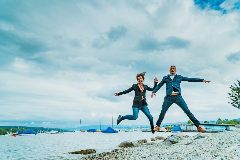 A man and woman jumping joyfully on a pebbly beach.