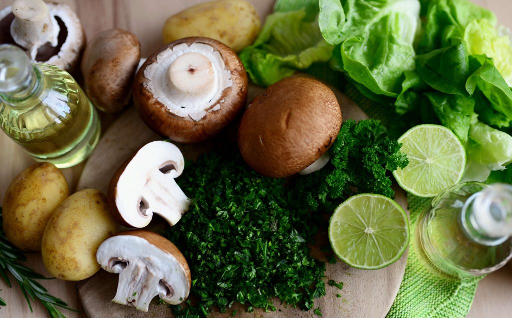Fresh whole ingredients including mushrooms, potatoes, lime, and leafy greens on a wooden cutting board